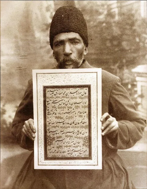صورة فوتوغرافية لميرزا محمد رضا كلهر بقلنسوة من فرو الحمل، يحمل لوحة خطية مؤطّرة